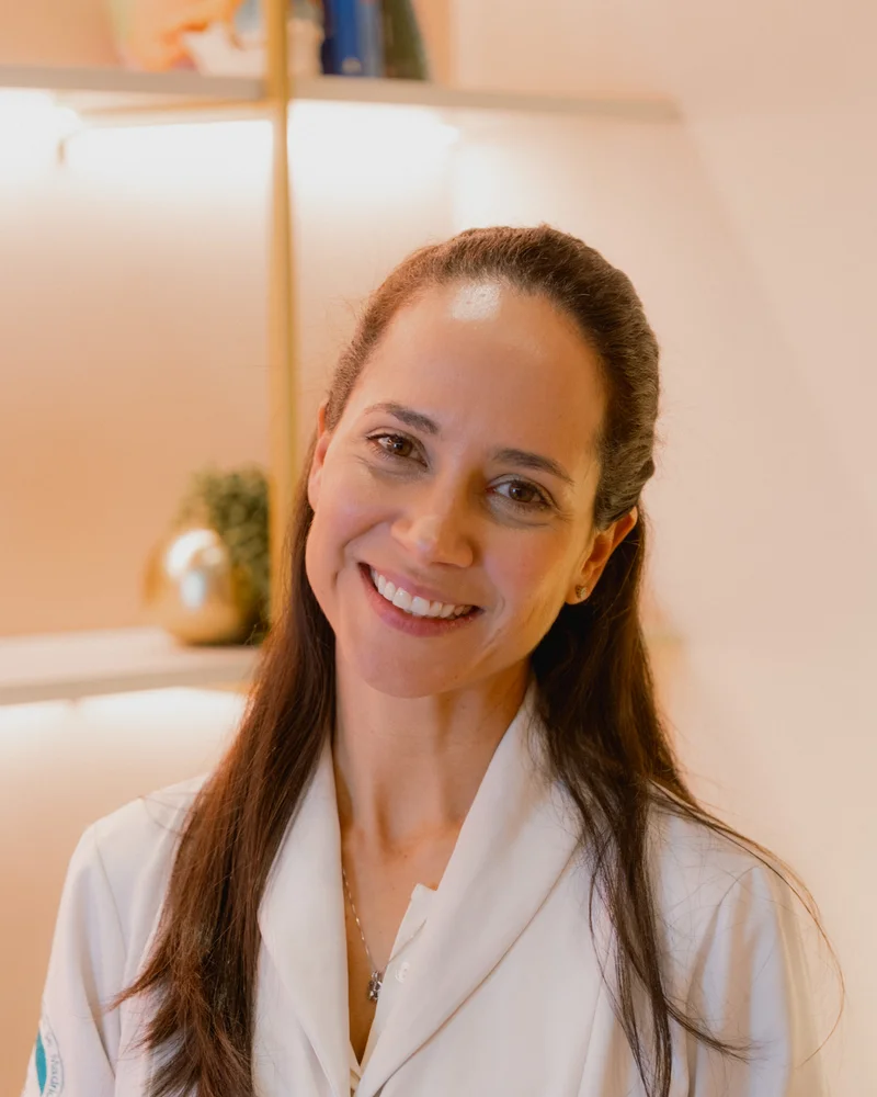 Dra. Laura Proença sorrindo em jaleco, fundo da Clínica Laura Proença